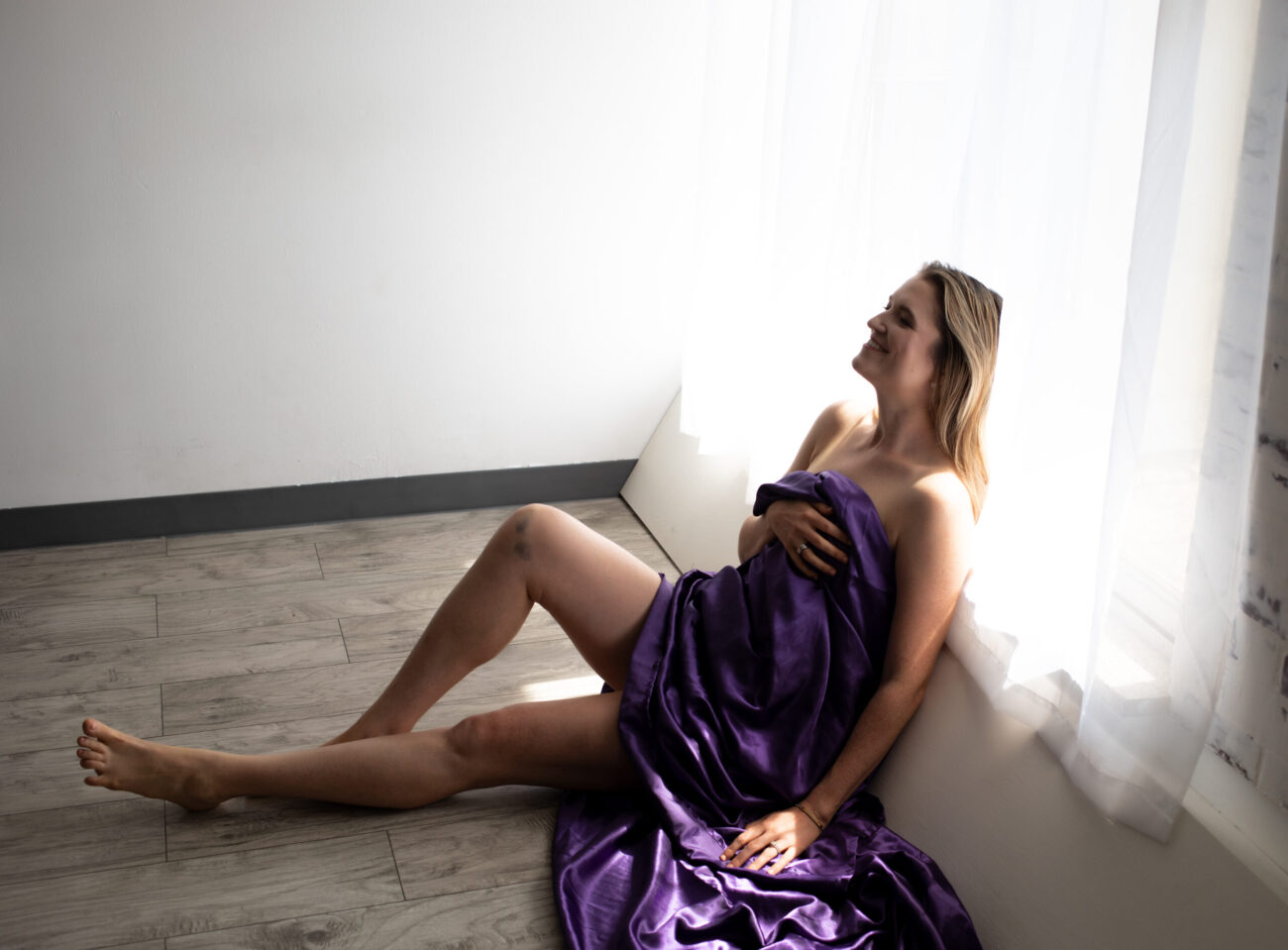 Brunette uses a purple sheet to cover while sitting and smiling against a window.